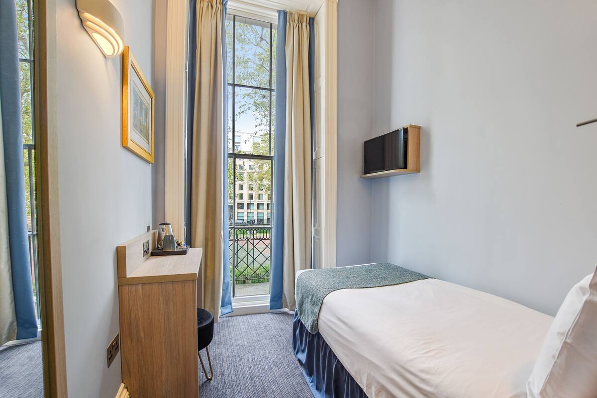 A hotel room with a bed, a desk with a chair, a TV mounted on the wall, and a window with curtains.