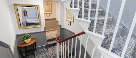 Un escalier avec des balustrades blanches et une main courante en bois, des marches recouvertes de moquette, une petite table avec une plante et un tableau encadré au mur.