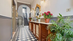A hallway with a checkered floor, a wooden console table with potted plants, a large ornate mirror, and a staircase in the background. - Judd Hotel (London)