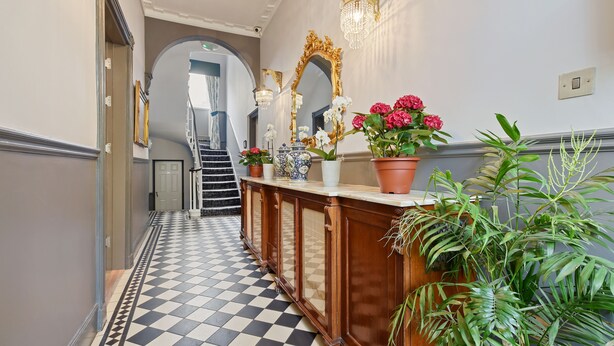 A hallway with a checkered floor, a wooden console table with potted plants, a large ornate mirror, and a staircase in the background.