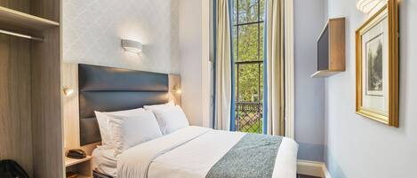 A hotel room with a bed, a wardrobe, a bedside table with a lamp, a window with curtains, and a framed picture on the wall.