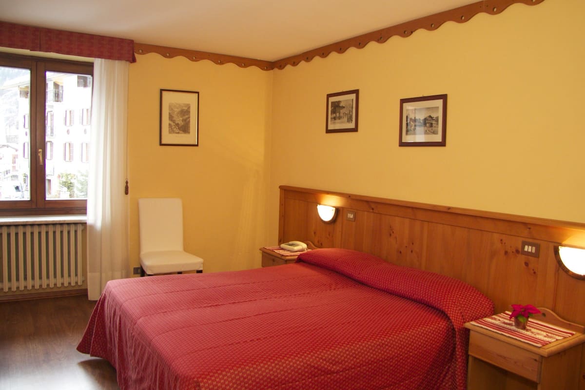 Habitación de hotel con cama, una silla, mesita de noche con lámpara y cuadros en la pared.