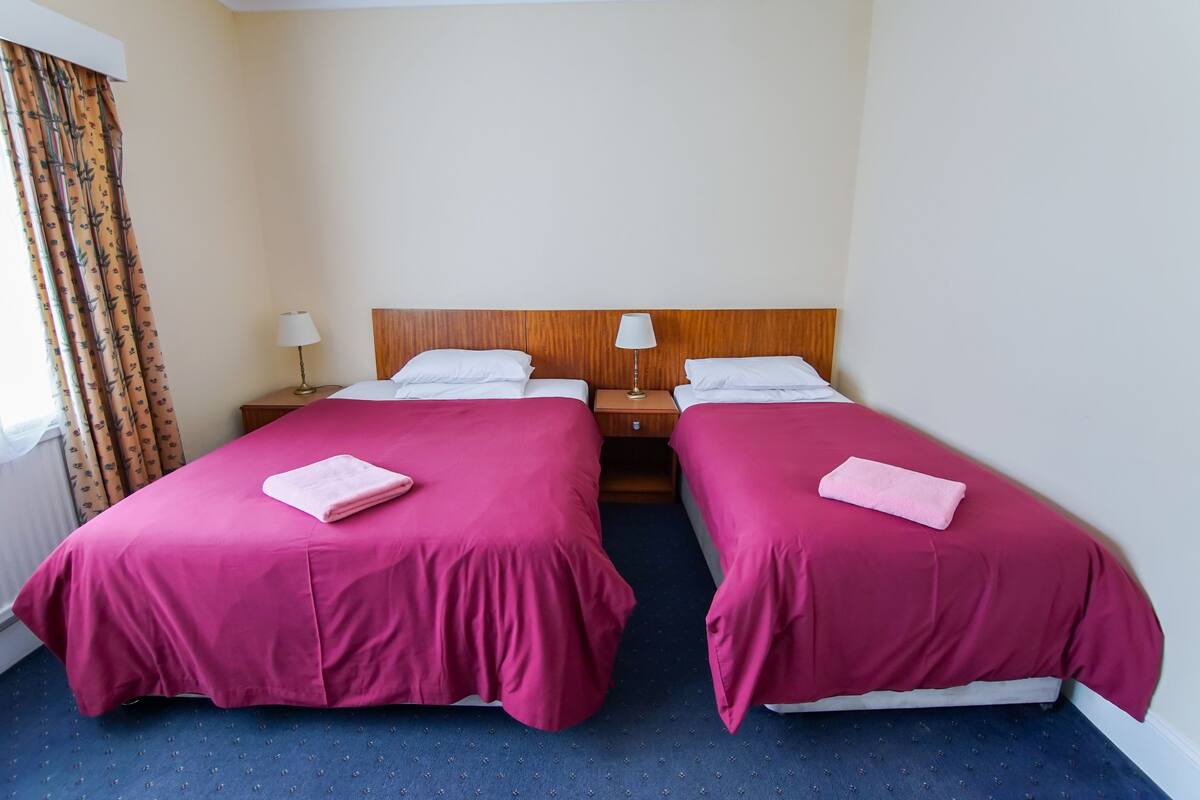 A hotel room with two beds, each with a pink bedspread and a white pillow. There is a wooden headboard, a nightstand with a lamp, and a window with curtains.