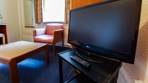A hotel room with a large flat-screen TV on a stand, a wooden table, a pink chair, and a small black table with a remote control.