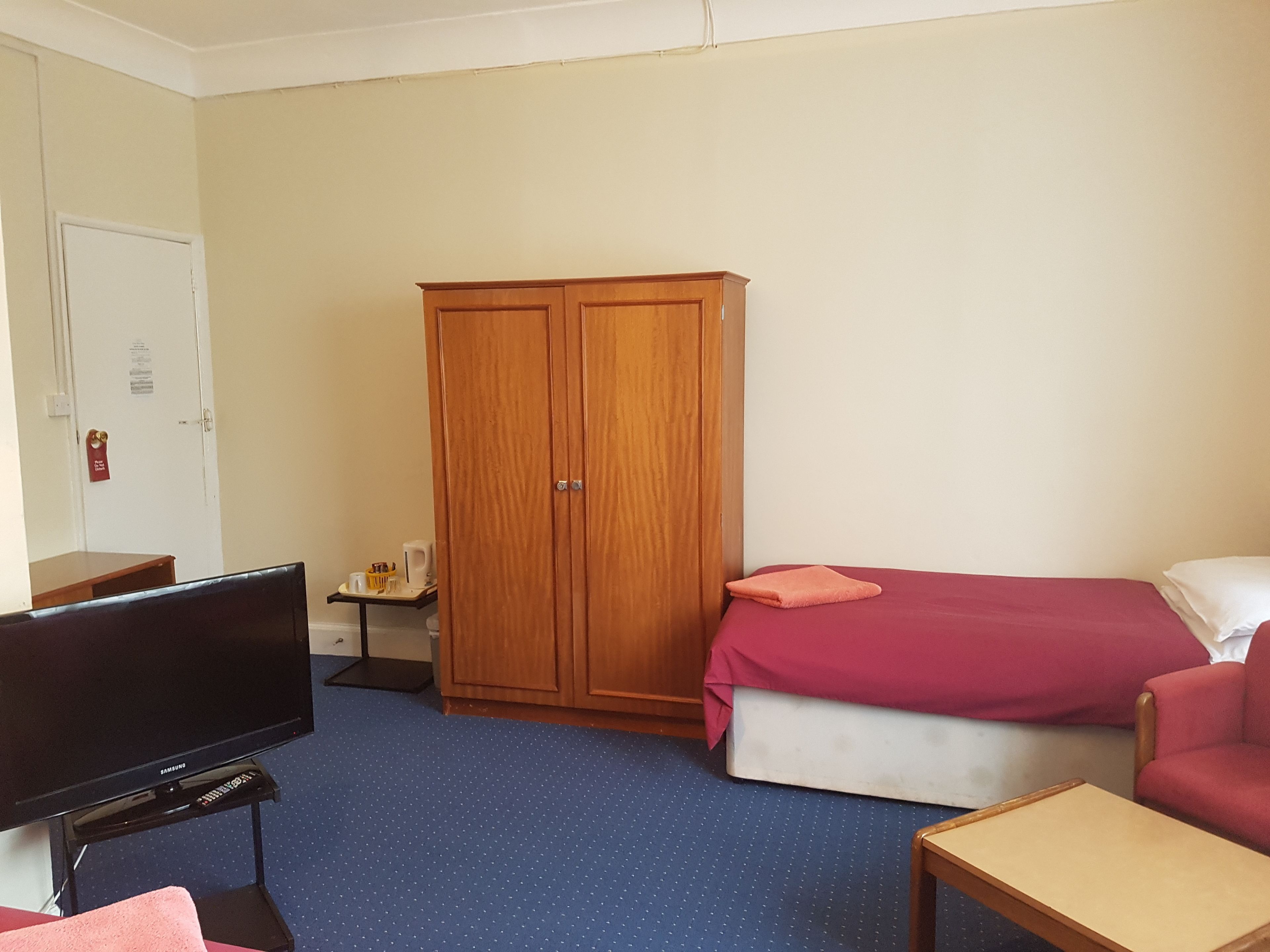 Una habitación de hotel con una cama, un televisor sobre un mueble, un armario de madera y una pequeña mesa con una silla.