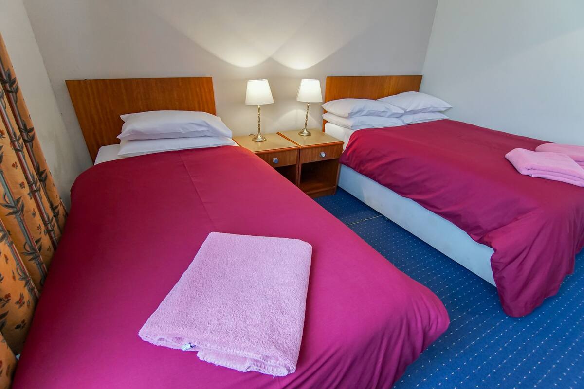 A hotel room with two beds, each with a red bedspread and white pillows. There are two bedside tables with lamps, and a folded towel on the left bed.