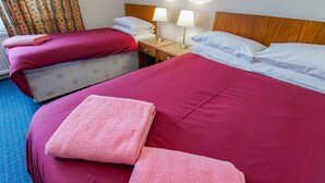 A hotel room with two beds, each with red bedding and white pillows. There are two bedside tables with lamps, and a blue carpeted floor.