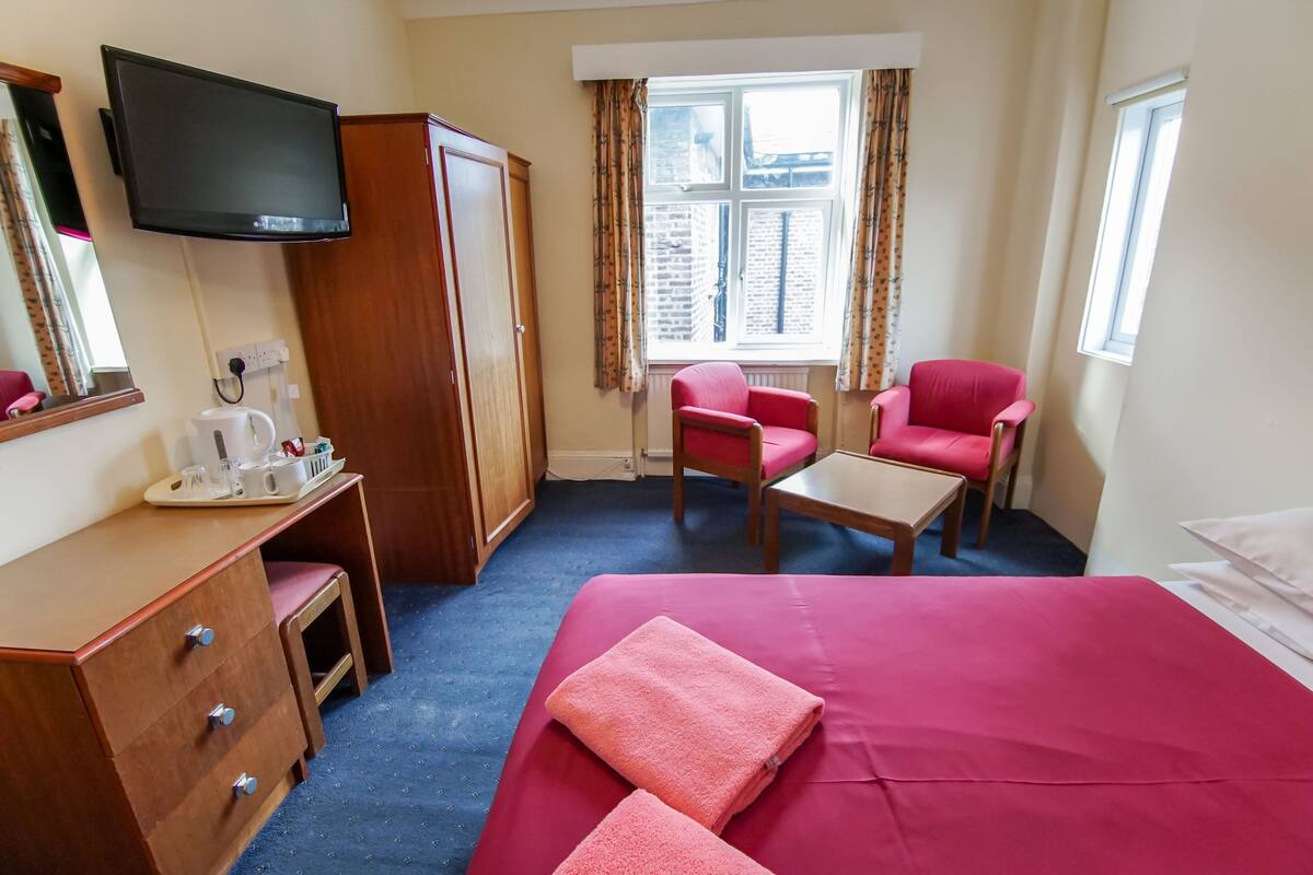 A hotel room with a bed, two chairs, a desk, a television, and a window with curtains.