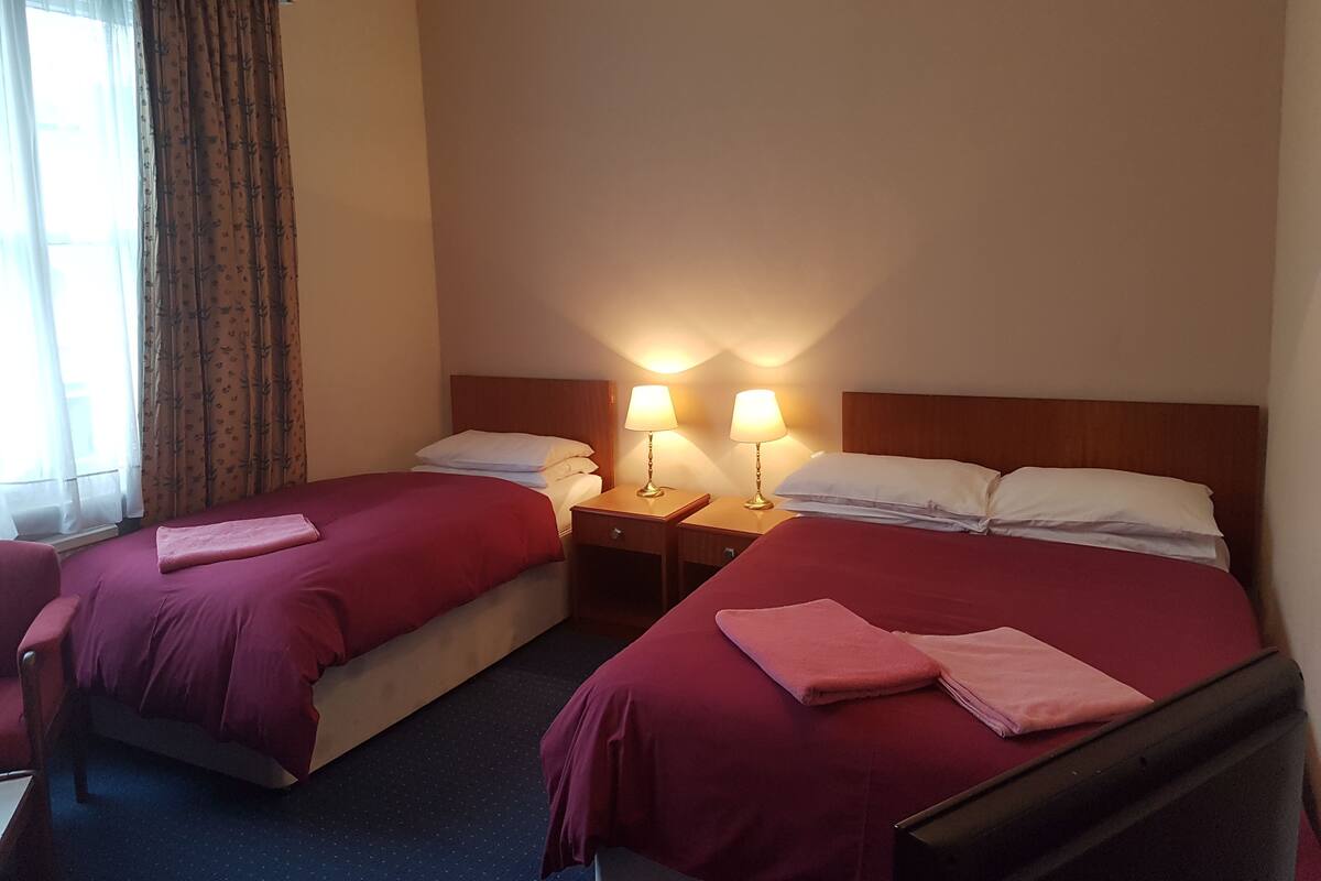 A hotel room with two beds, each with a red bedspread and white pillows. There are two bedside lamps and a window with curtains.