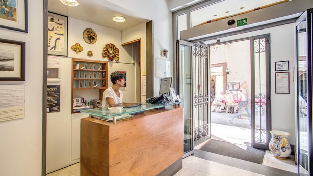 A reception desk with a wooden finish and glass top, flanked by framed artwork and a shelf with decorative items. - Hotel Nuova Italia (Florence)