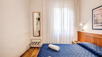 Ein Schlafzimmer mit einem hölzernen Kopfteil, einer blau gesteppten Bettdecke, zwei aufgerollten Handtüchern, einem Nachttisch mit Lampe und einem Fenster mit transparenten Vorhängen.
