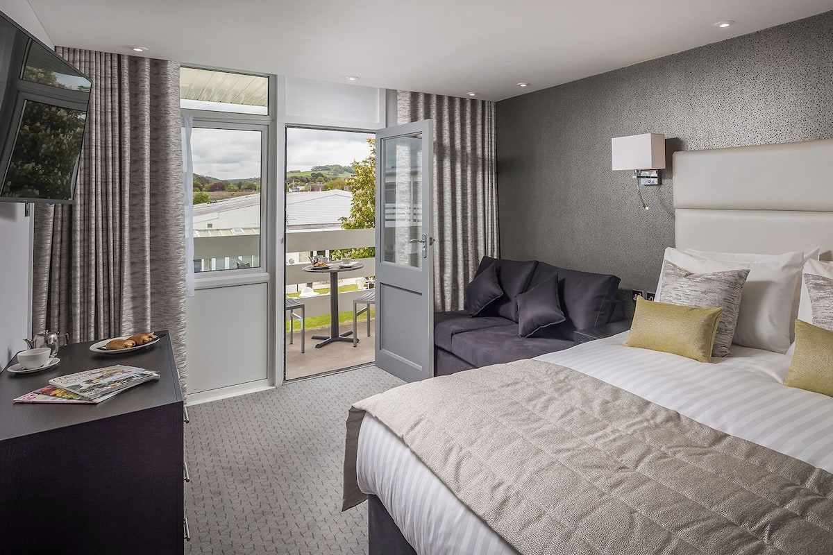 A modern hotel room with a large bed, a desk, and a balcony with a view of the countryside.