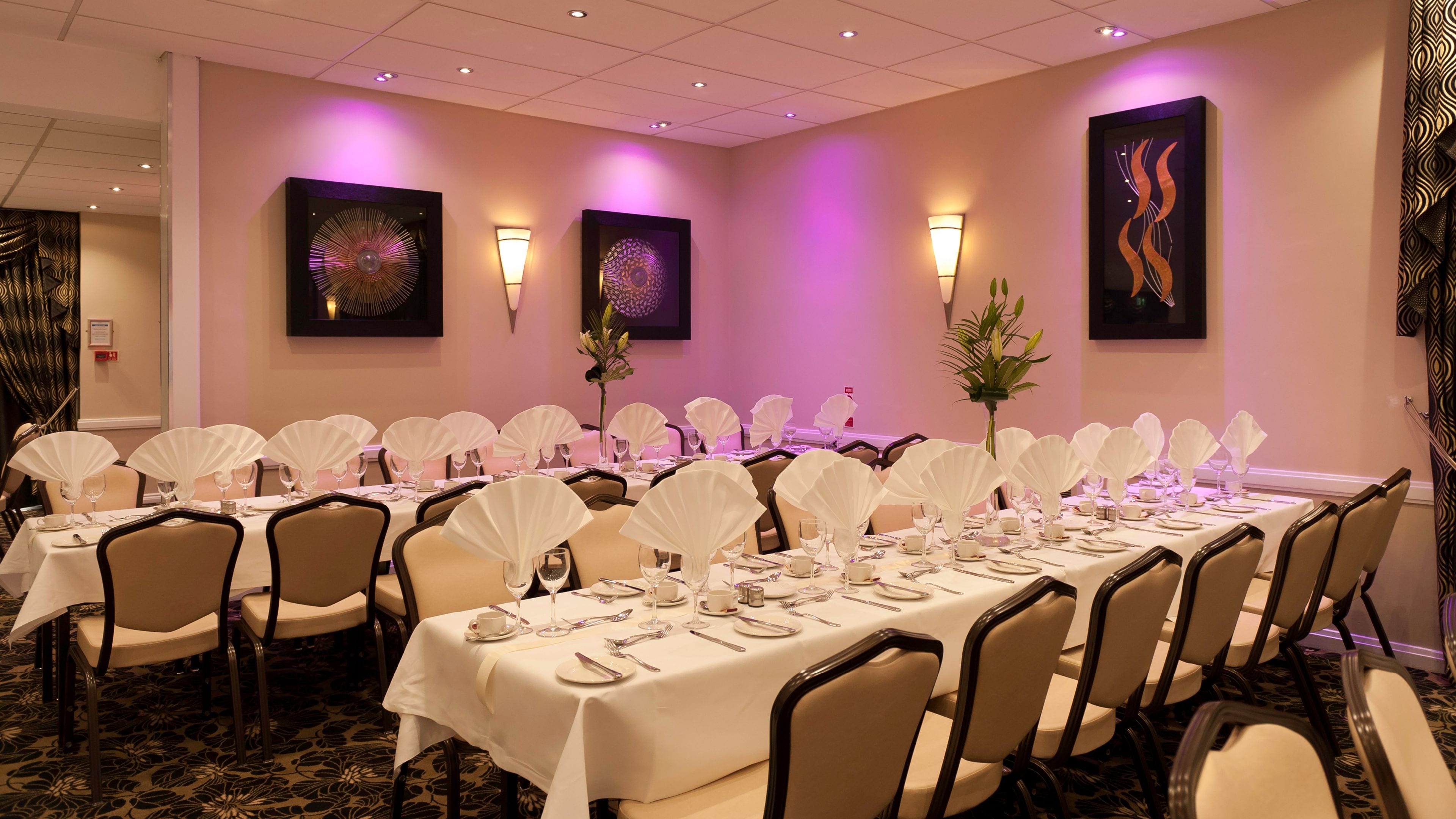 A conference room with tables set for a formal event, featuring white tablecloths, chairs, and decorative wall art.