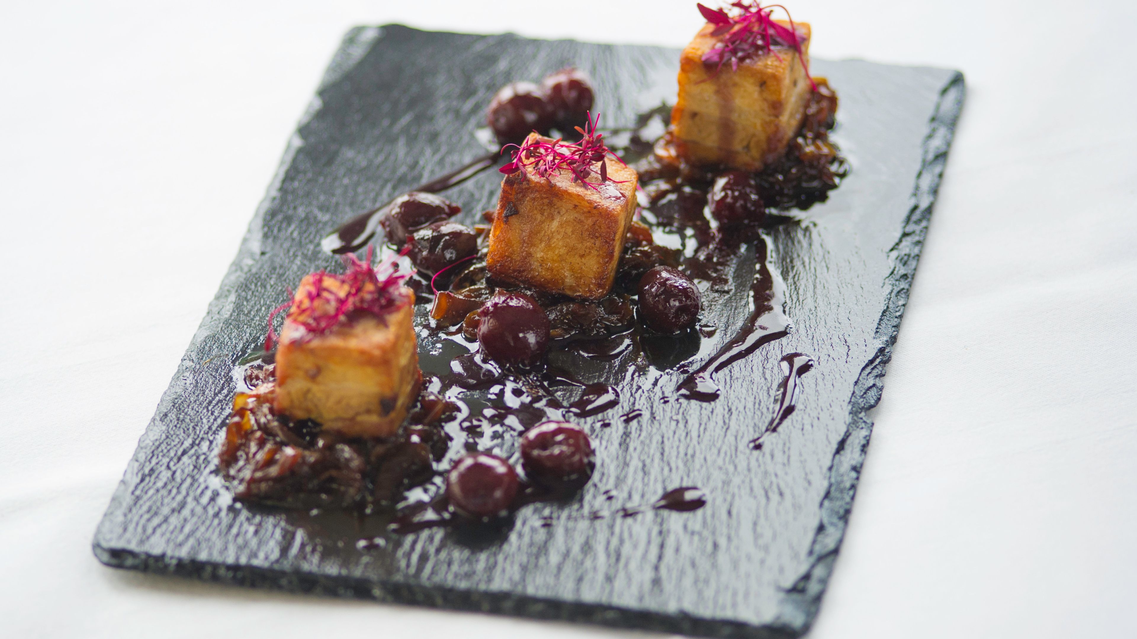 Plato rectangular de pizarra con tres porciones de comida cuadrada, adornado con flores comestibles rojas, rodeado de salsa oscura y bayas.