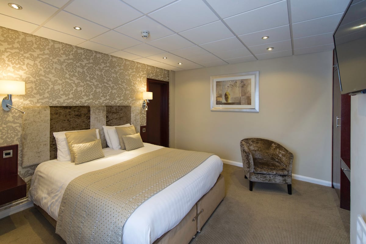 A hotel room with a large bed, two wall-mounted lamps, a framed artwork, a chair, and a telephone on the nightstand.