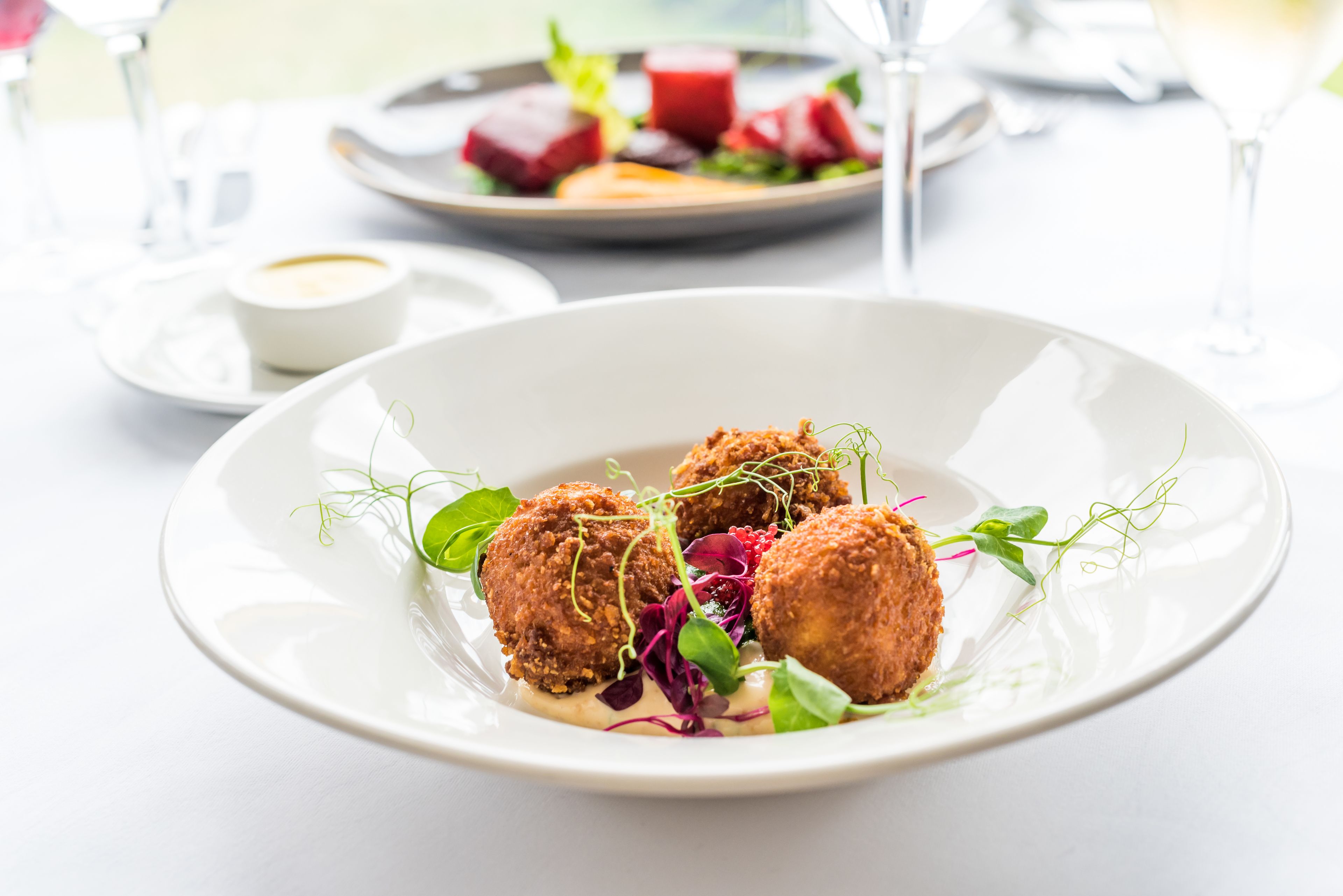Une assiette blanche contenant trois aliments frits et des légumes verts, un petit bol de sauce et un verre de vin blanc, le tout posé sur une table.