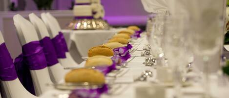 Una mesa de comedor formal con sillas blancas, cintas moradas y una fila de postres.