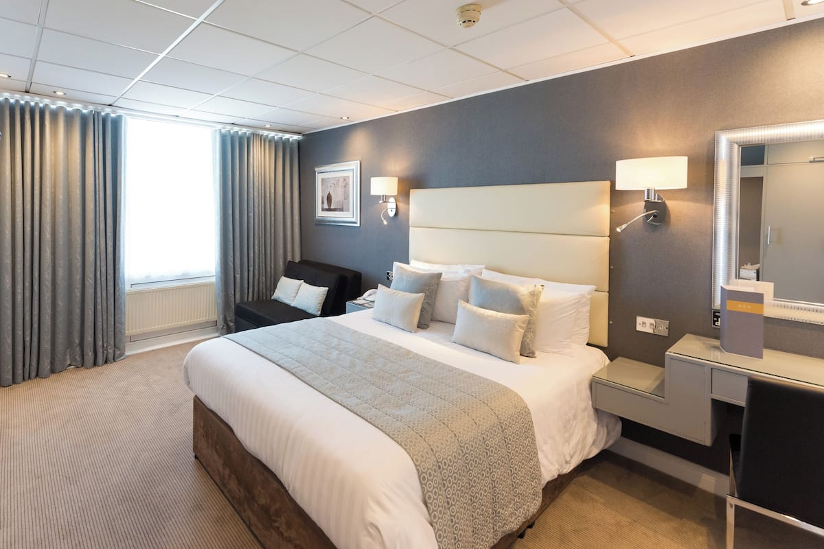 A hotel room with a large bed, a bedside table, a desk, a chair, a flat-screen TV, a window with curtains, and a wall-mounted lamp.
