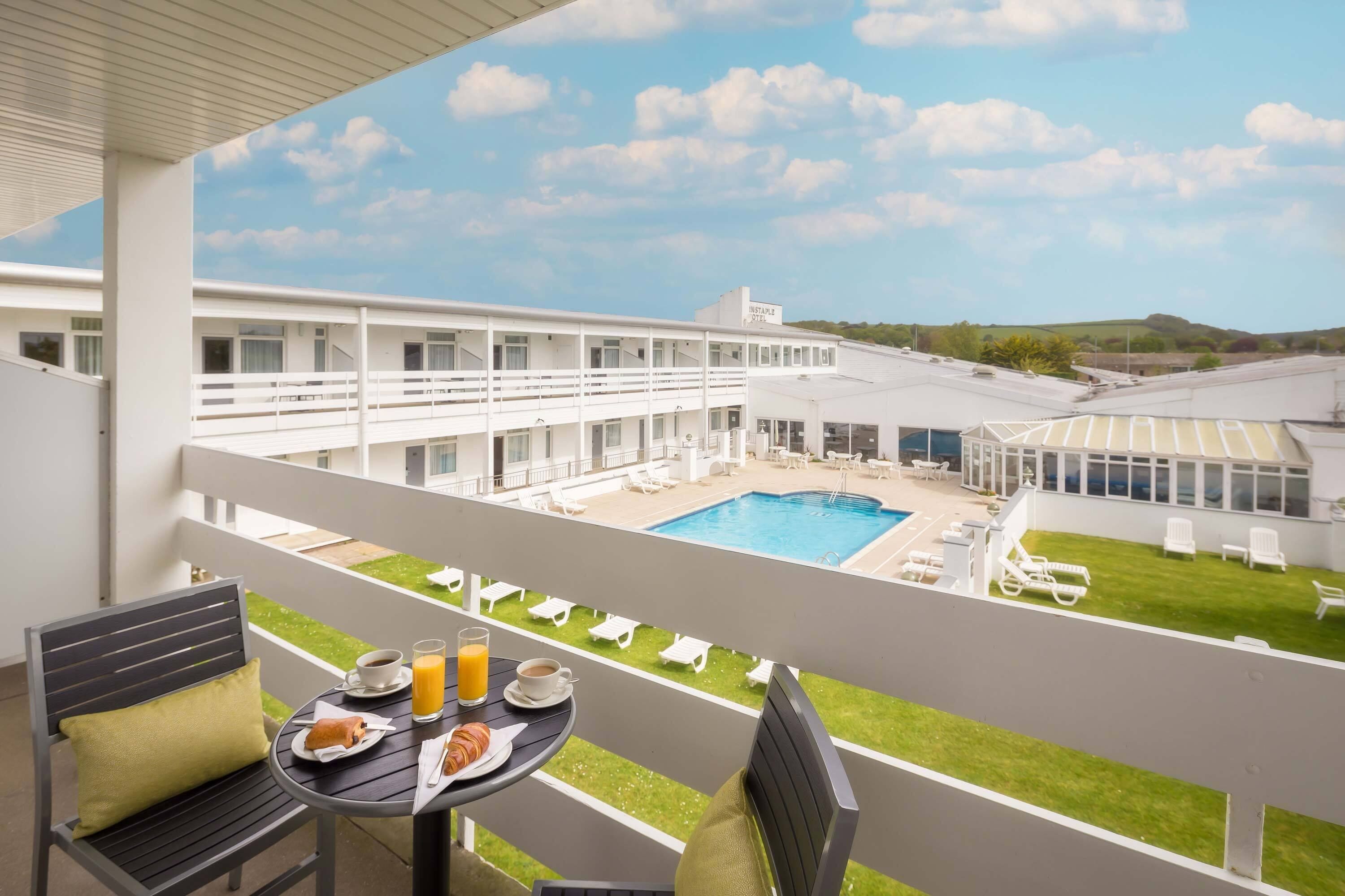 A balcony with a table set for breakfast, overlooking a pool area with lounge chairs and a building with a sign that reads "Resort."