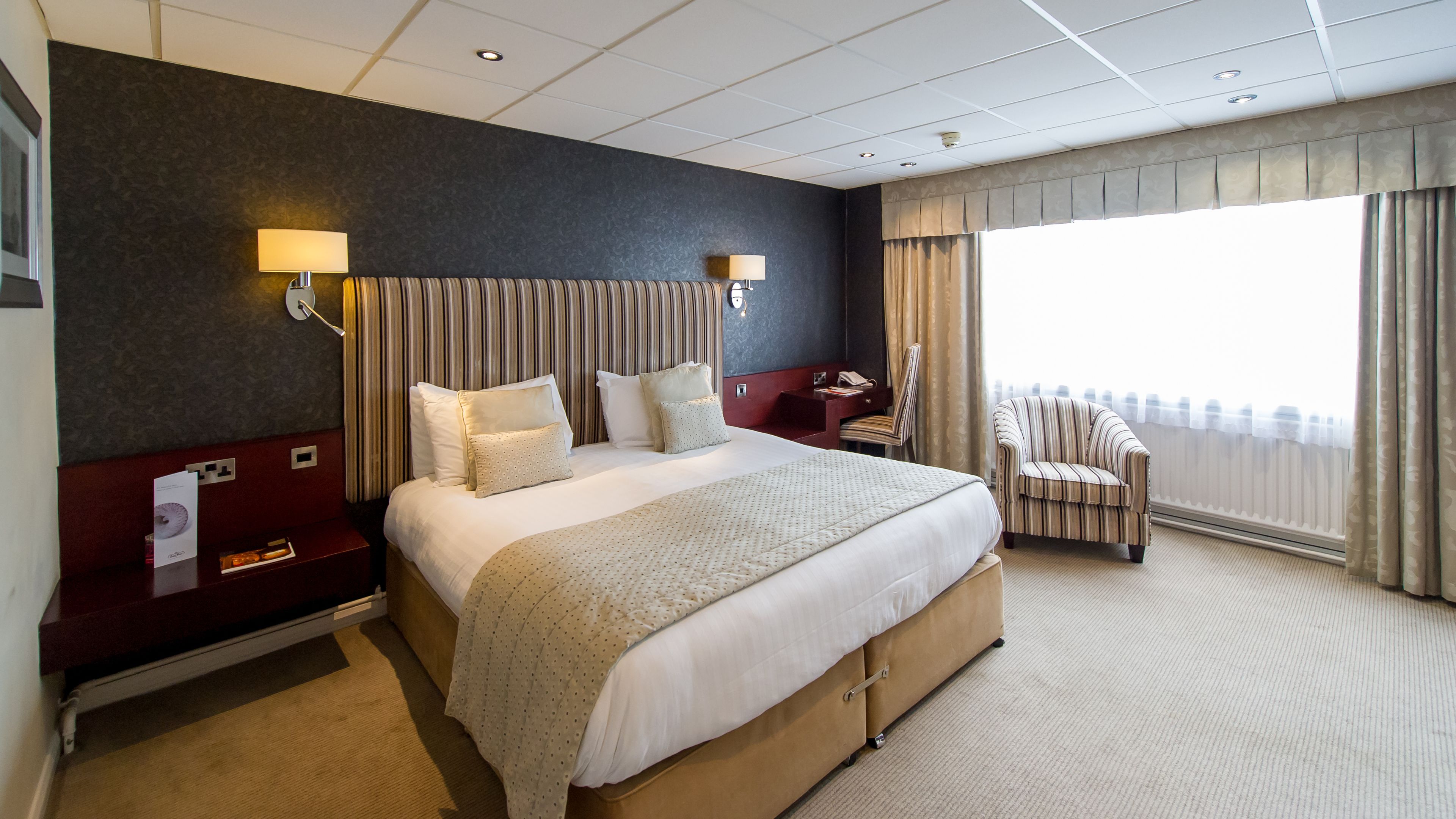 A hotel room with a large bed, two bedside lamps, a chair, a desk, and a large window with curtains.