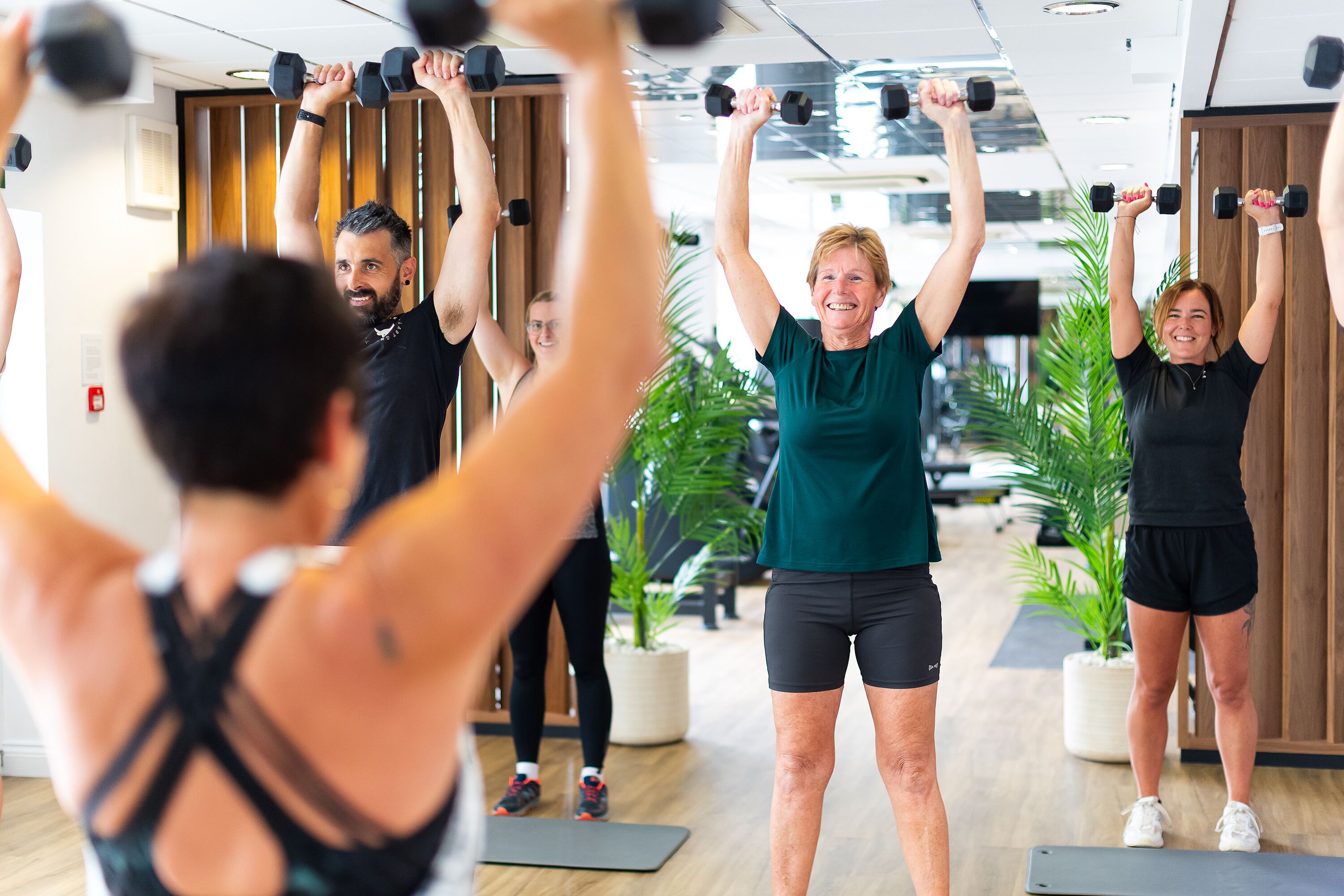 A group of people exercising with dumbbells in a room with wooden panels and potted plants.