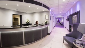 A reception area with a curved counter, a receptionist, a chair, and a hallway leading to a health club. - The Barnstaple Hotel (Barnstaple)