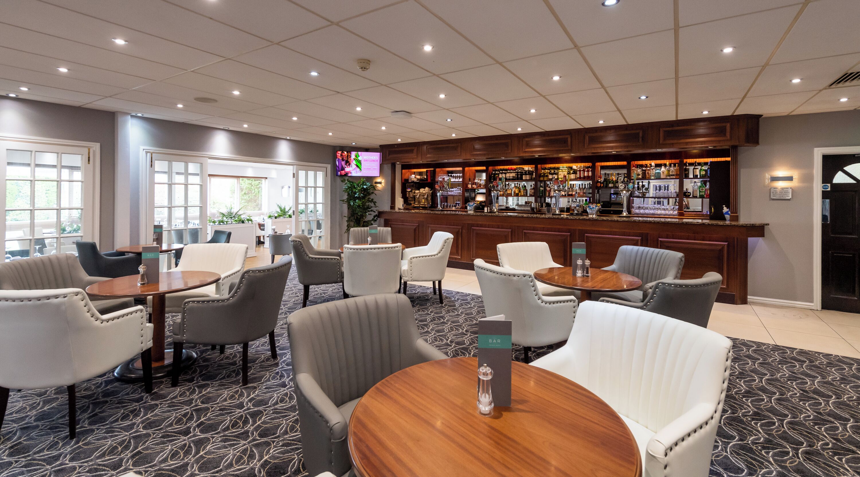 Un hall d’hôtel avec des tables et des chaises rondes, un coin bar où sont exposées différentes bouteilles, et une grande fenêtre laissant entrer la lumière naturelle.