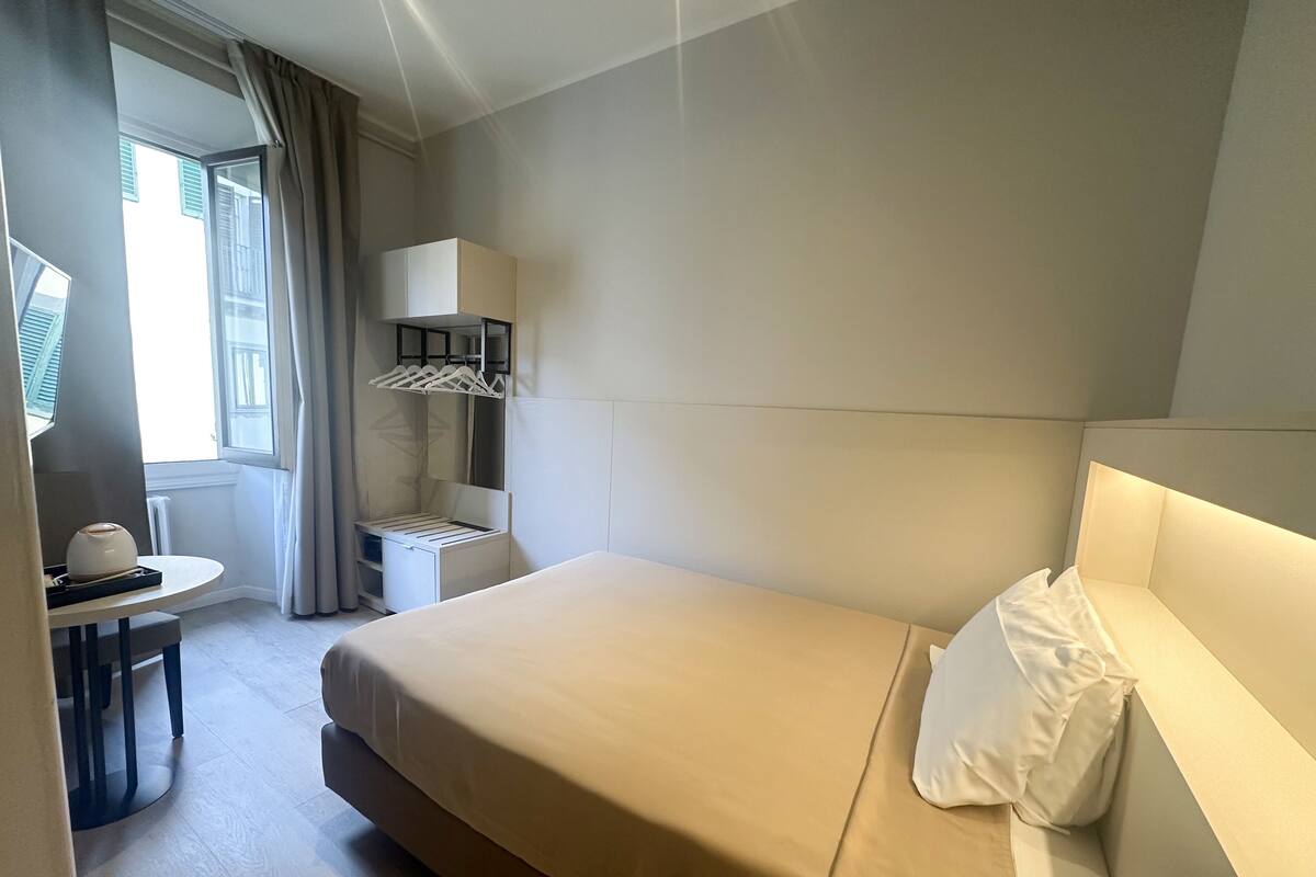 A hotel room with a bed, a bedside table, a mirror, a wardrobe, and a window with curtains.