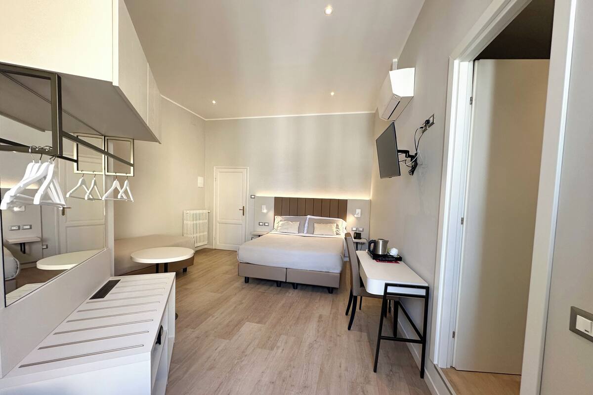 A modern hotel room with a bed, bedside table, and a small desk with a chair. There is a mirror, a radiator, and a wall-mounted TV.