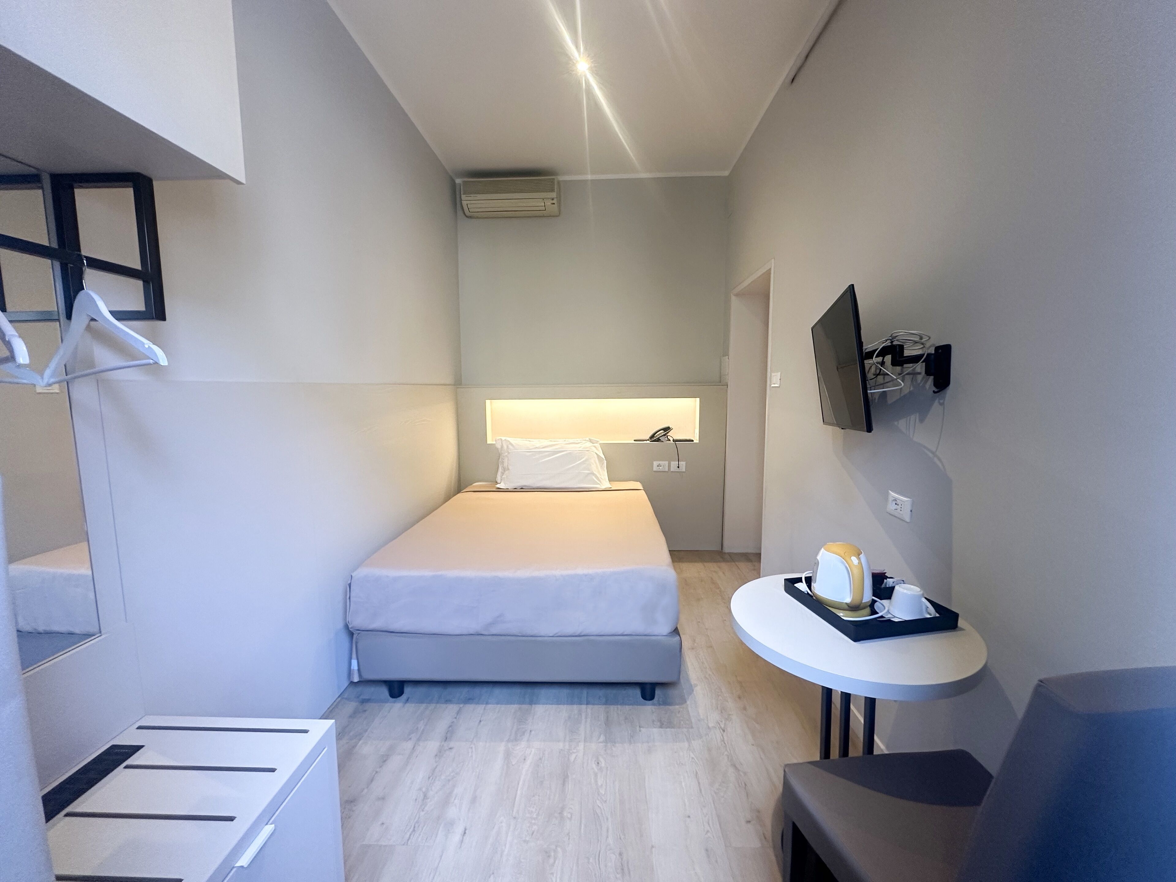 Ein modernes Hotelzimmer mit einem Einzelbett, einem kleinen Tisch mit Frühstücksgebäck und -speisen, einem an der Wand montierten Fernseher sowie einer Decken-Klimaanlage.