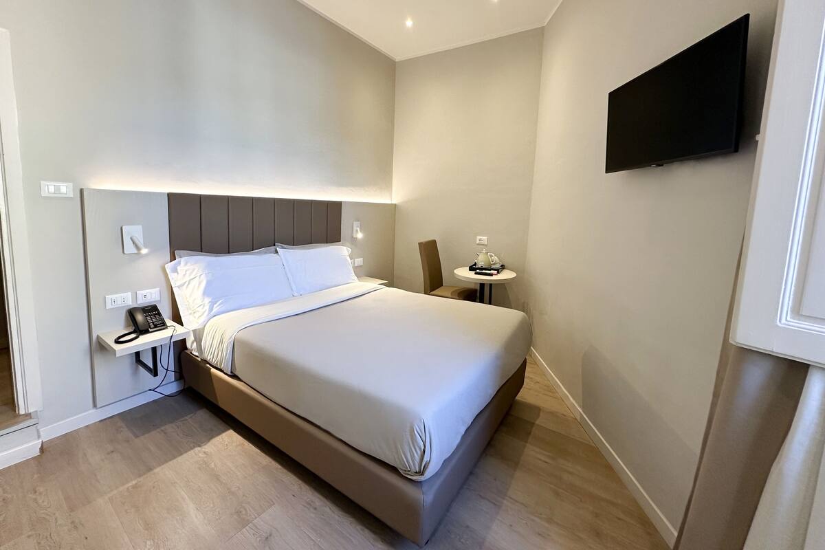 A hotel room with a bed, bedside table, lamp, and a mounted TV.