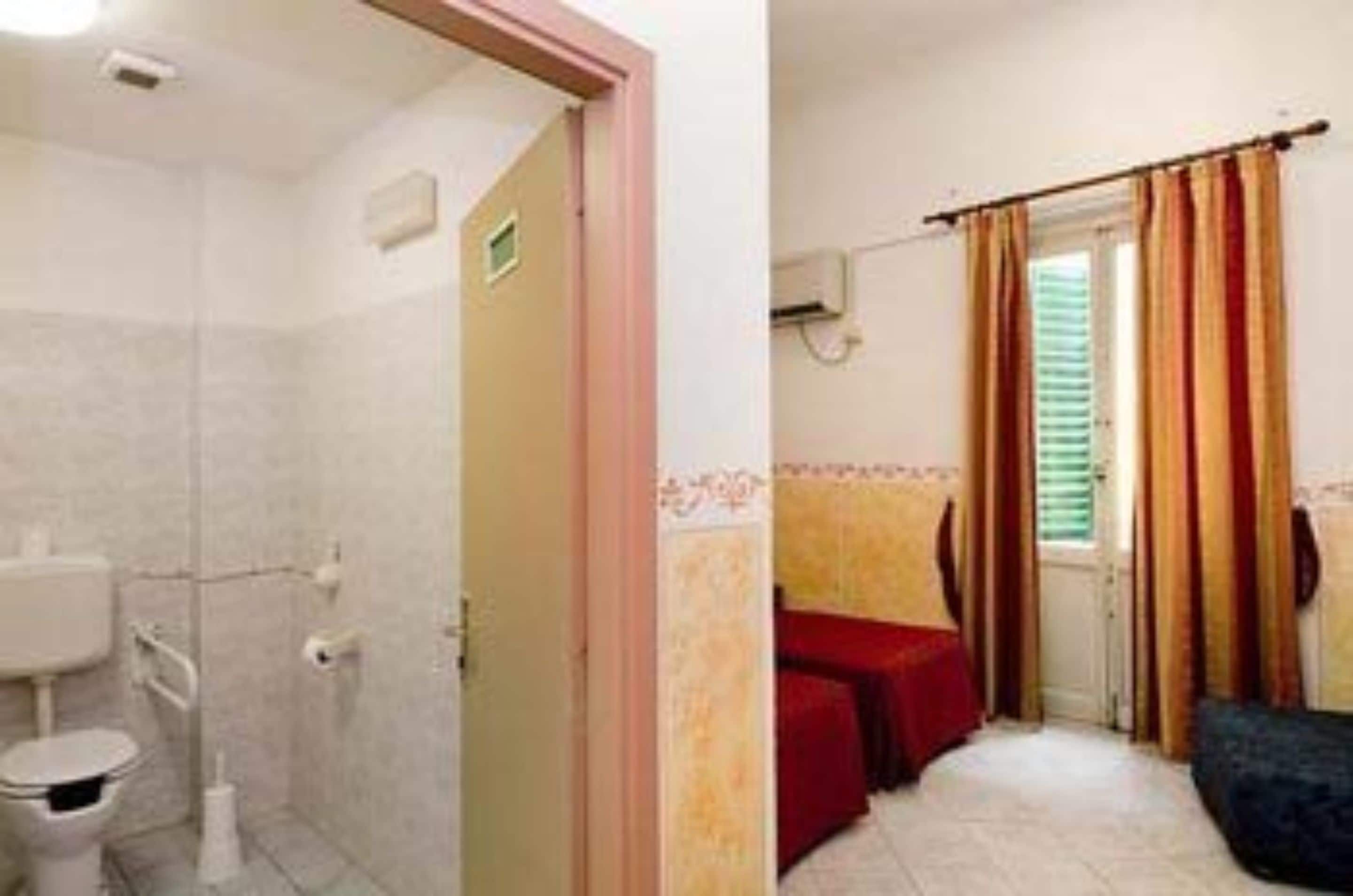 Une salle de bain avec des toilettes, un lavabo et une douche. Une porte donne sur une pièce où se trouvent un lit et des rideaux.