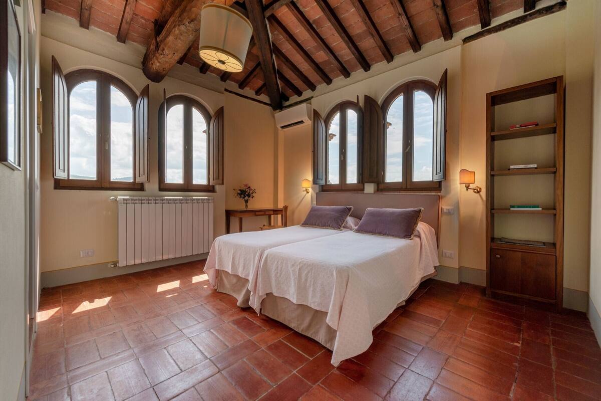 A bedroom with two beds, large arched windows, a radiator, and a wooden bookshelf.