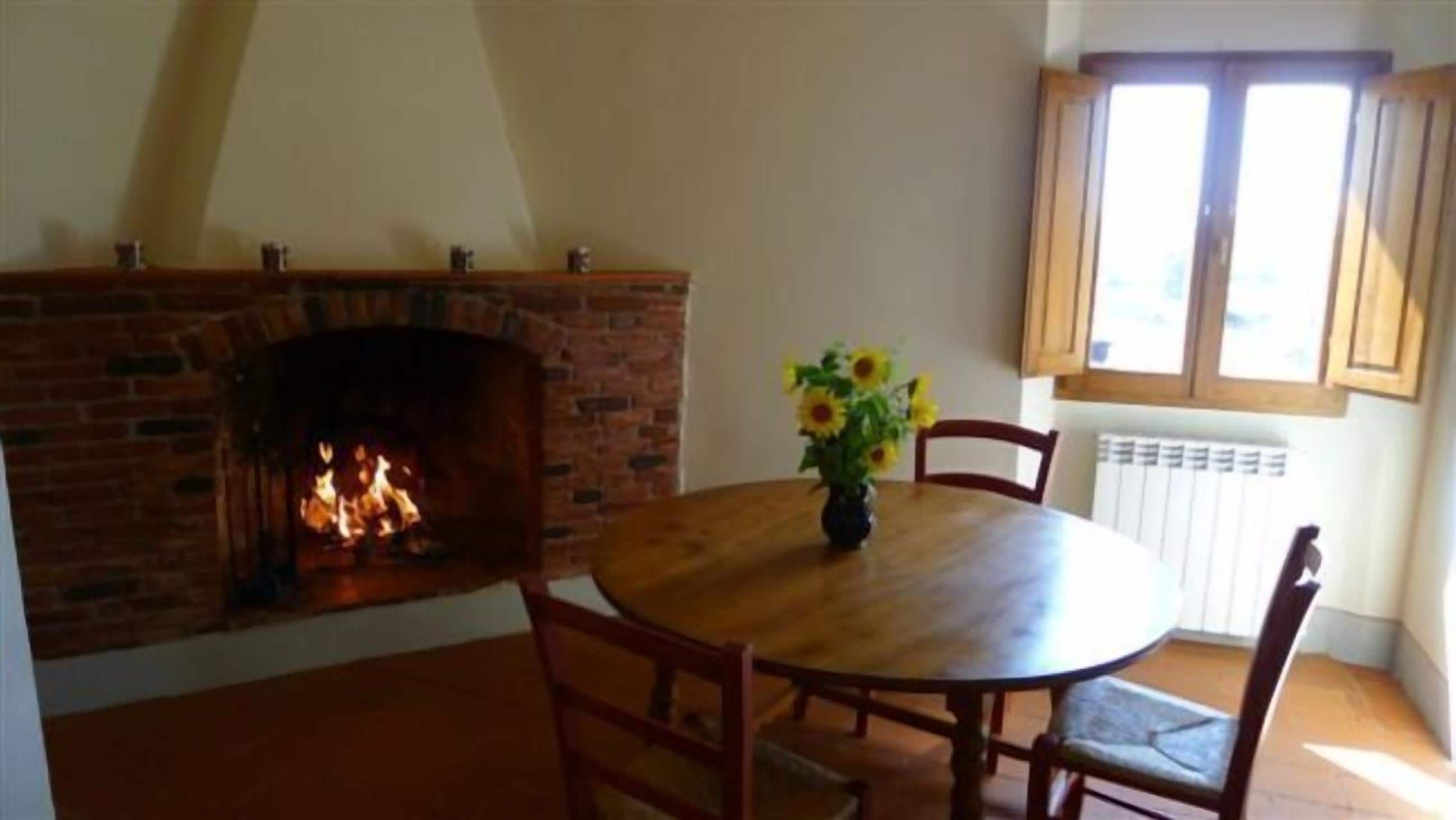 A room with a brick fireplace, a round wooden dining table with chairs, and a window with wooden shutters.
