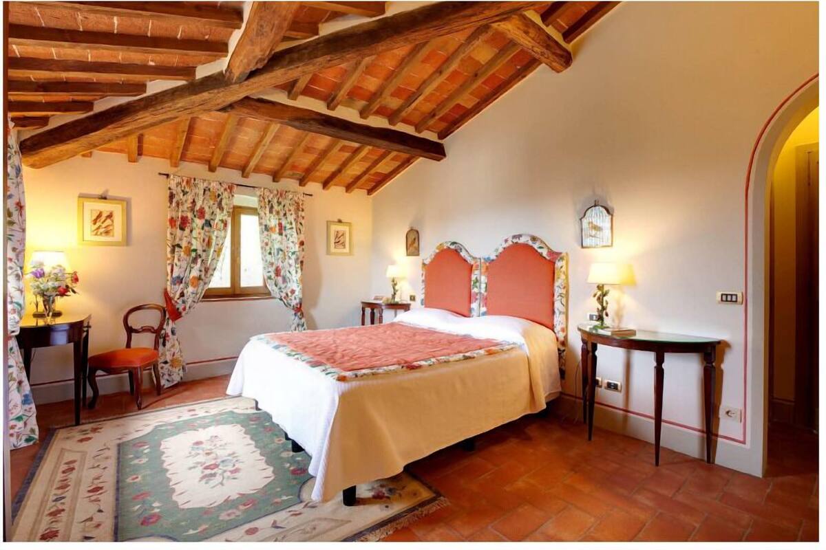 A bedroom with a wooden ceiling, a bed with a pink headboard, floral curtains, and a bedside table with a lamp.