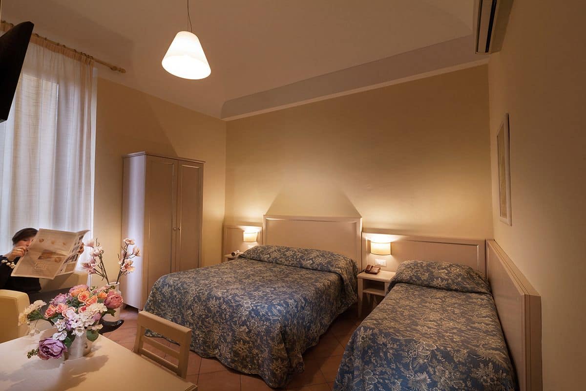 A hotel room with two beds, a desk, a chair, a cabinet, and a vase of flowers.