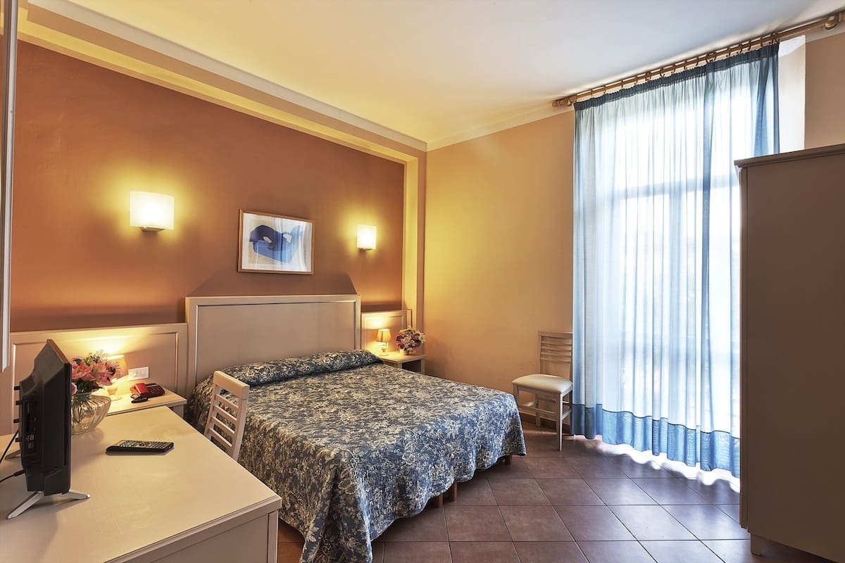 A hotel room with two beds, a desk with a computer and phone, a bedside table with a lamp and flowers, and a window with curtains.