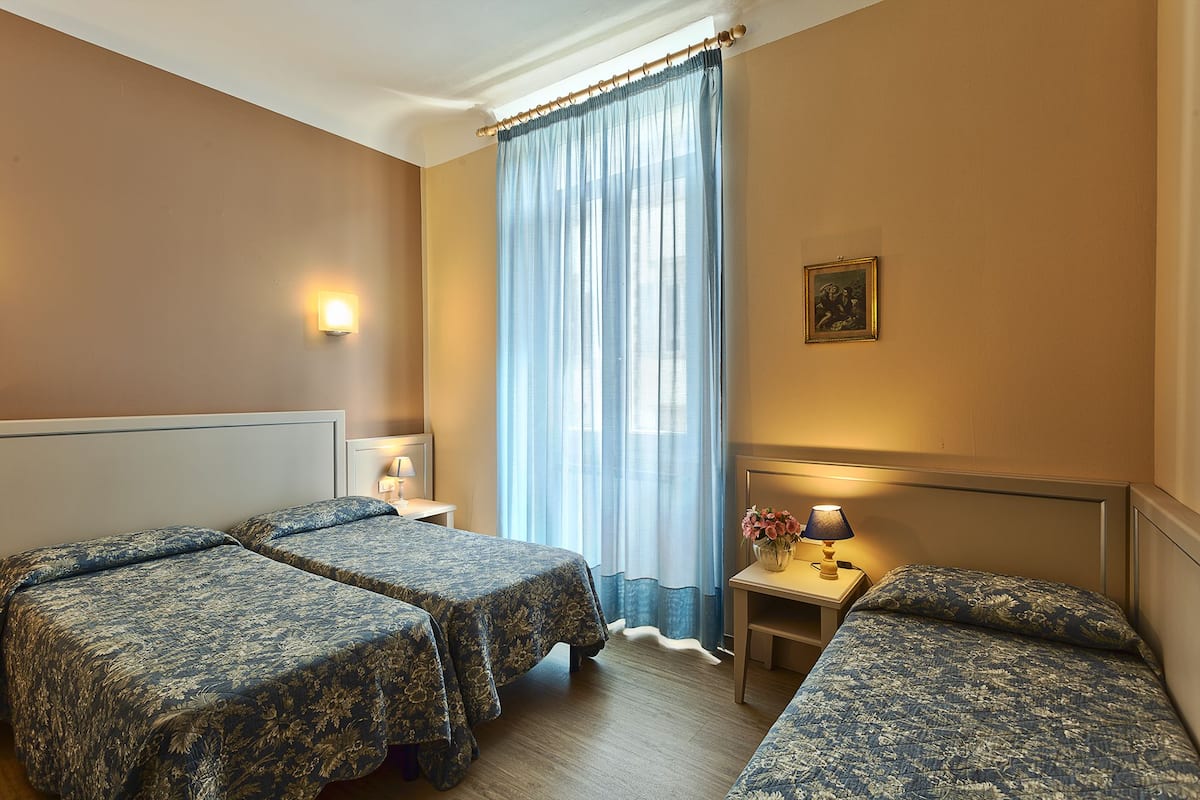 A hotel room with two beds, a nightstand with a lamp, a small table with a lamp, and a window with sheer curtains.