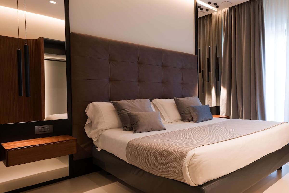 A modern bedroom with a large bed, a wooden nightstand, a mirror, and a view of the outdoors through a window with curtains.