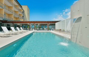 Une piscine avec des chaises longues, un espace de détente couvert et un bâtiment doté de balcons.