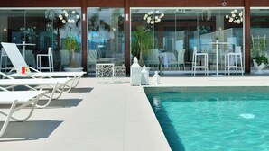 Un espace aménagé au bord de la piscine, avec des chaises longues et des tables, une piscine à eau claire et des portes vitrées donnant sur une salle à manger intérieure.