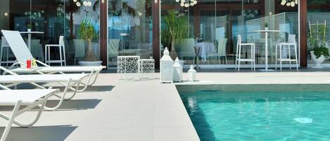 Zona junto a la piscina con sillones y mesas, una piscina cristalina y puertas de vidrio que dan a un comedor interior.
