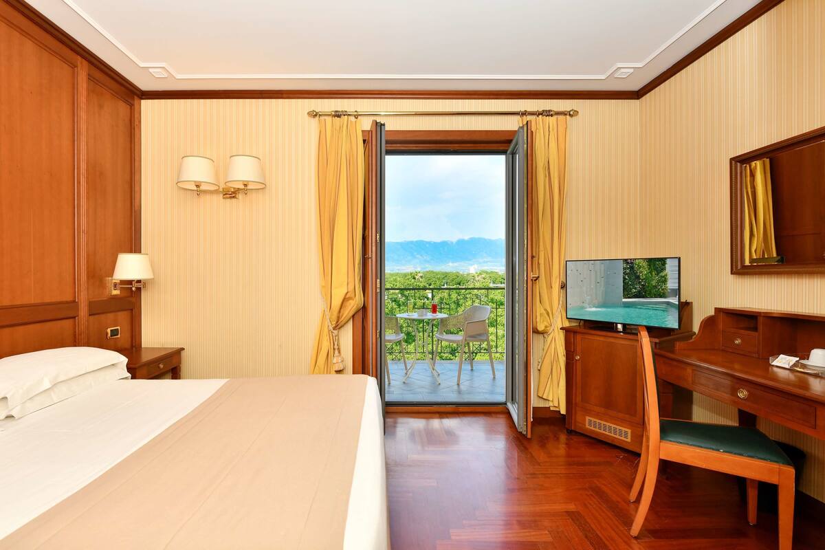 A hotel room with a bed, a desk, a television, and a balcony with a table and chairs.