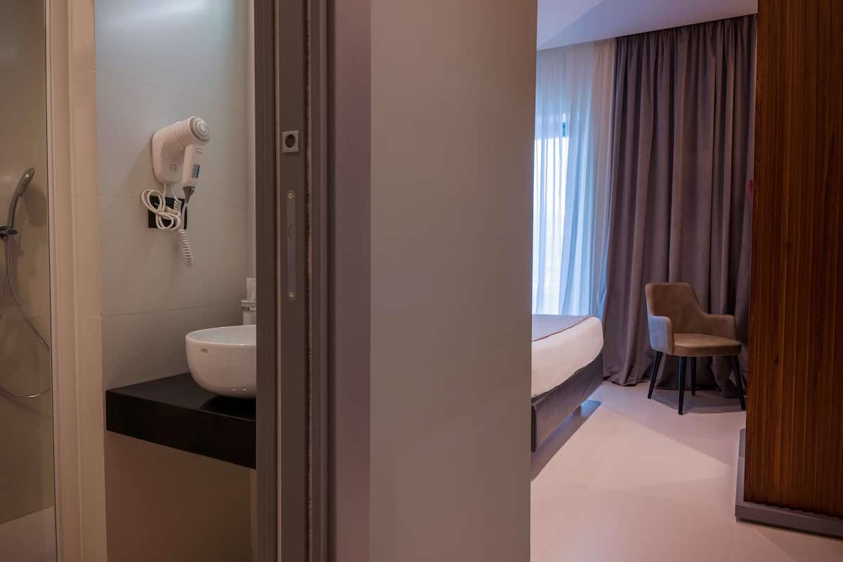A hotel bathroom with a hairdryer, a sink, and a shower. There is a bed with a chair in the adjacent room.