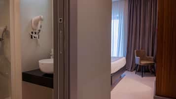 Un baño de hotel con secador de pelo, lavamanos y ducha. En la habitación contigua hay una cama y una silla.