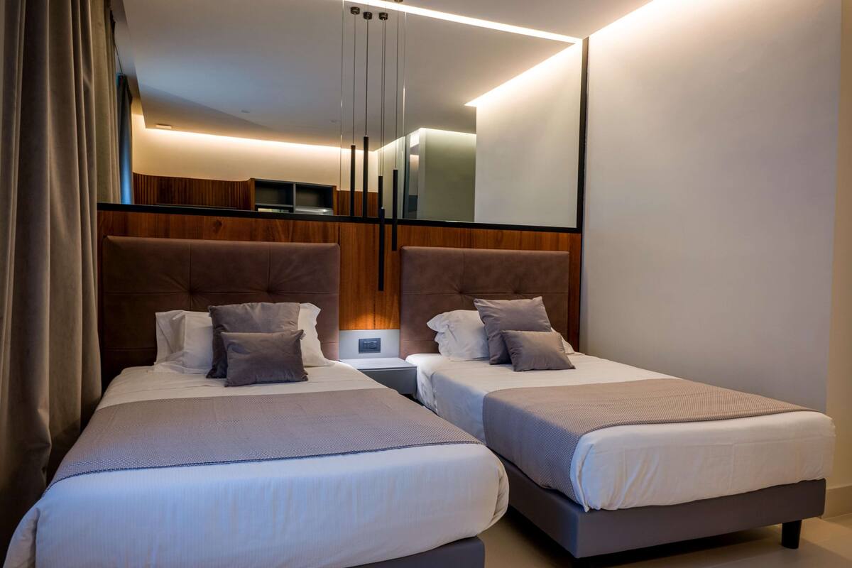 A modern hotel room with two beds, a wooden headboard, a bedside table, and a large mirror.