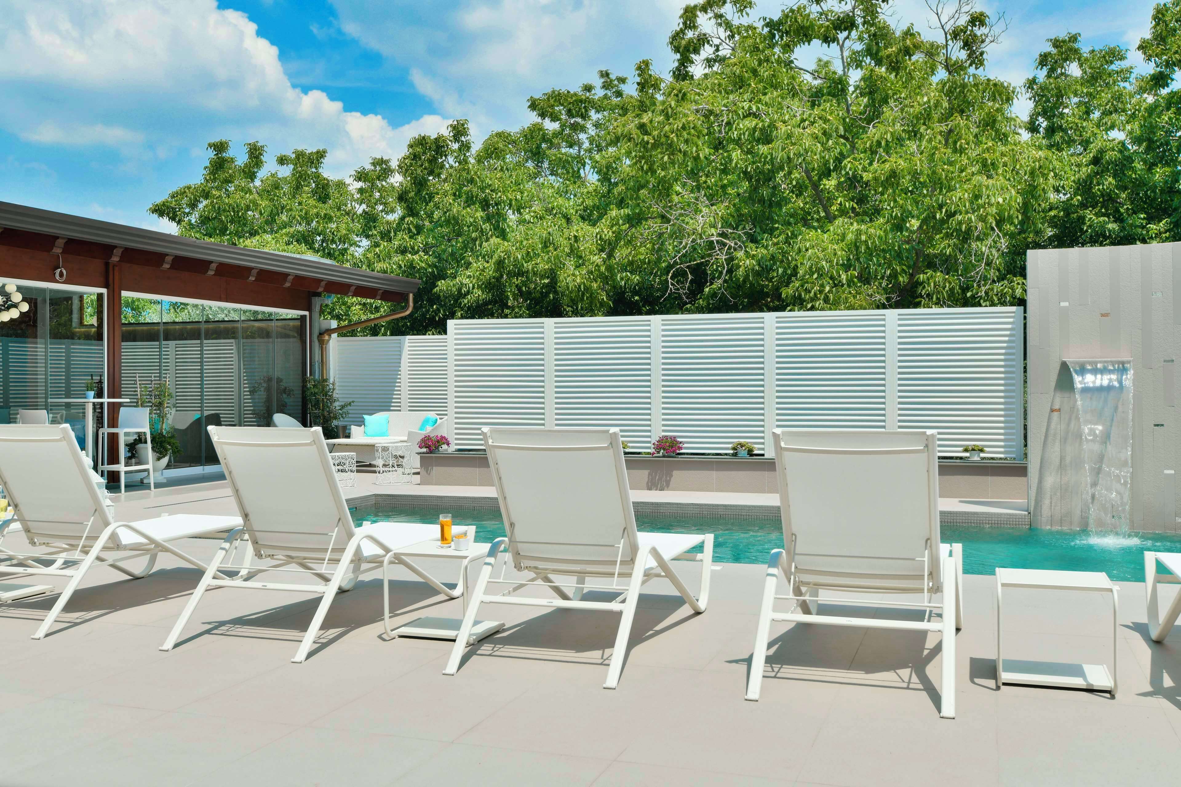 Zona de piscina con sillas reclinables blancas y una estructura de vidrio.