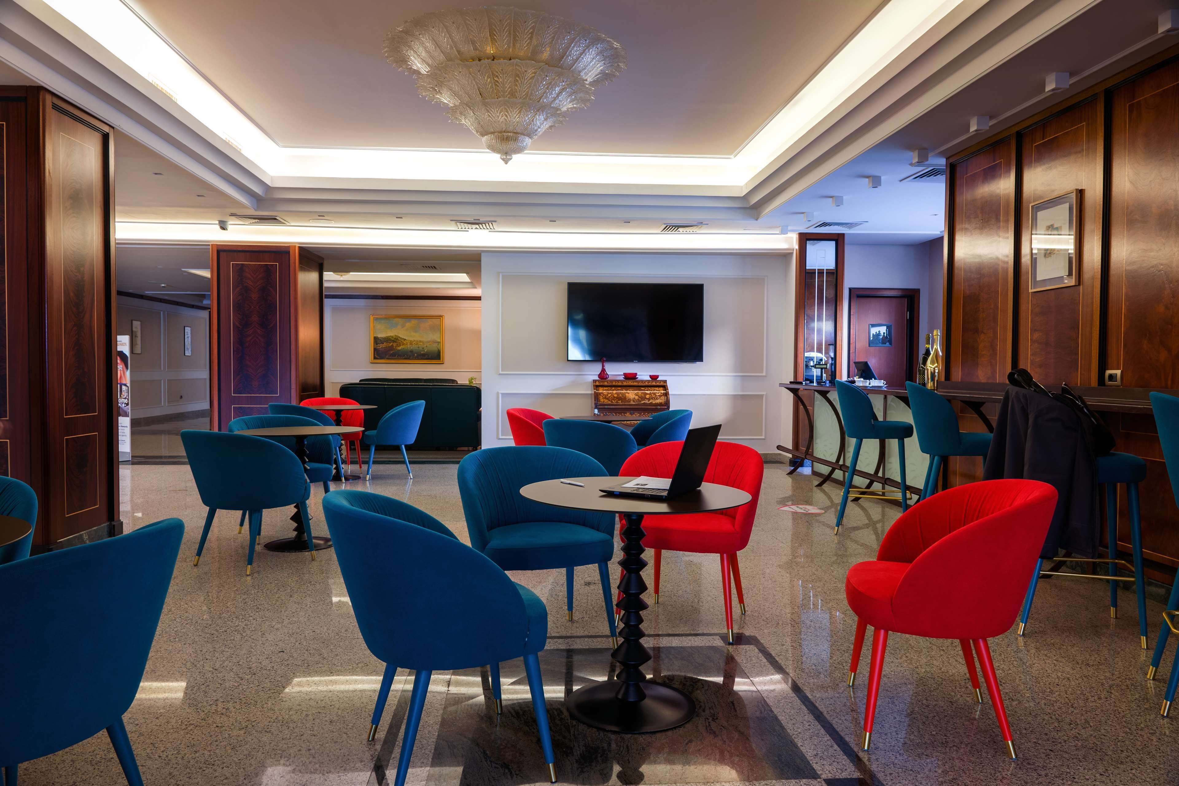 A modern lounge area with blue and red chairs, a round table with a laptop, and a large flat-screen TV on the wall.