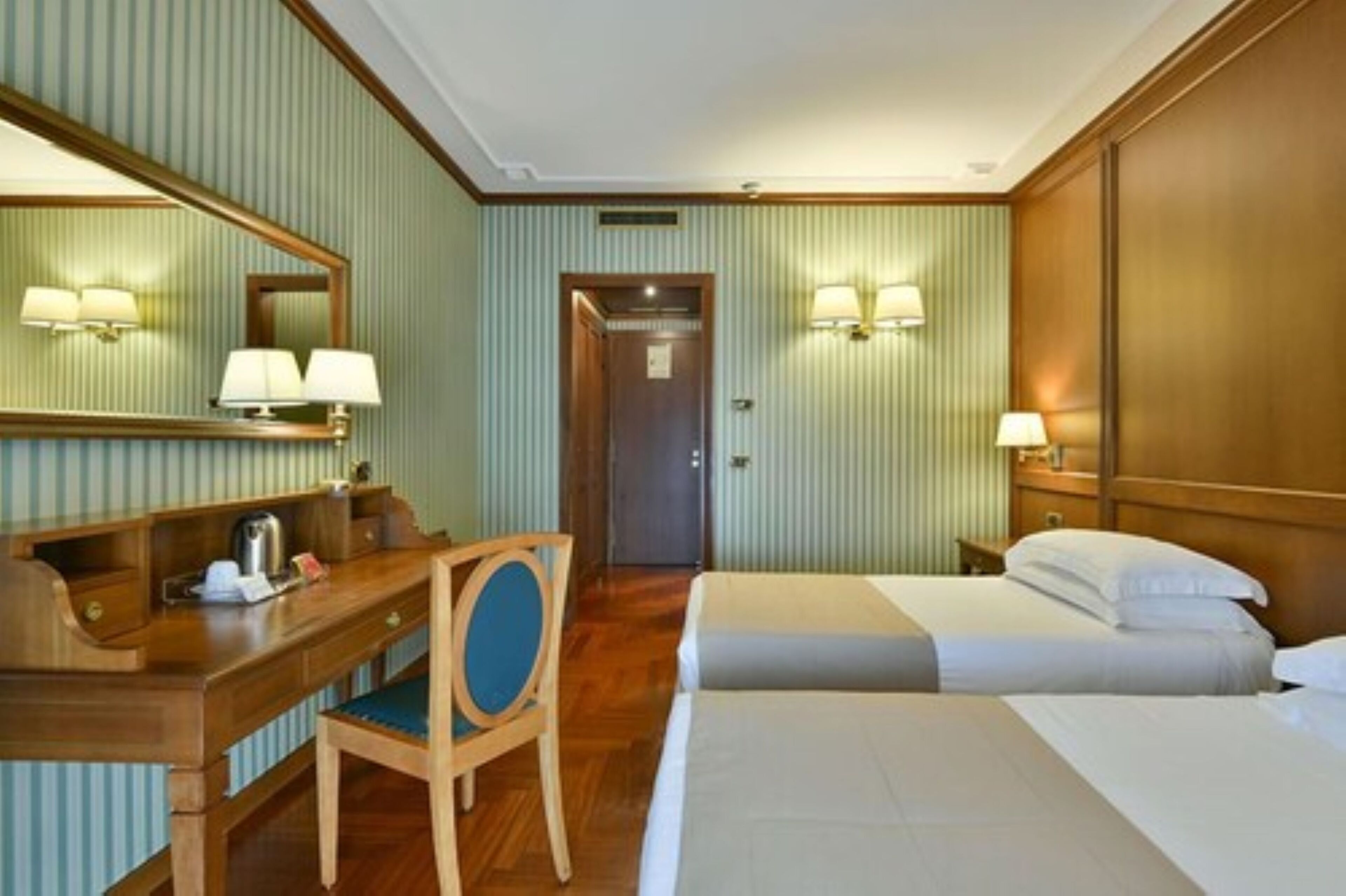 A hotel room with a wooden desk, two beds, a mirror, and wall-mounted lamps.