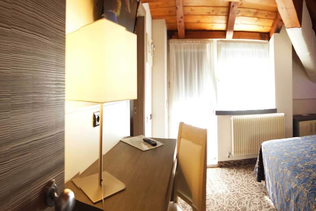Een hotelkamer met een houten plafond, een bed met een beddegoed met dessin, een nachtkastje met een lamp en een radiator.